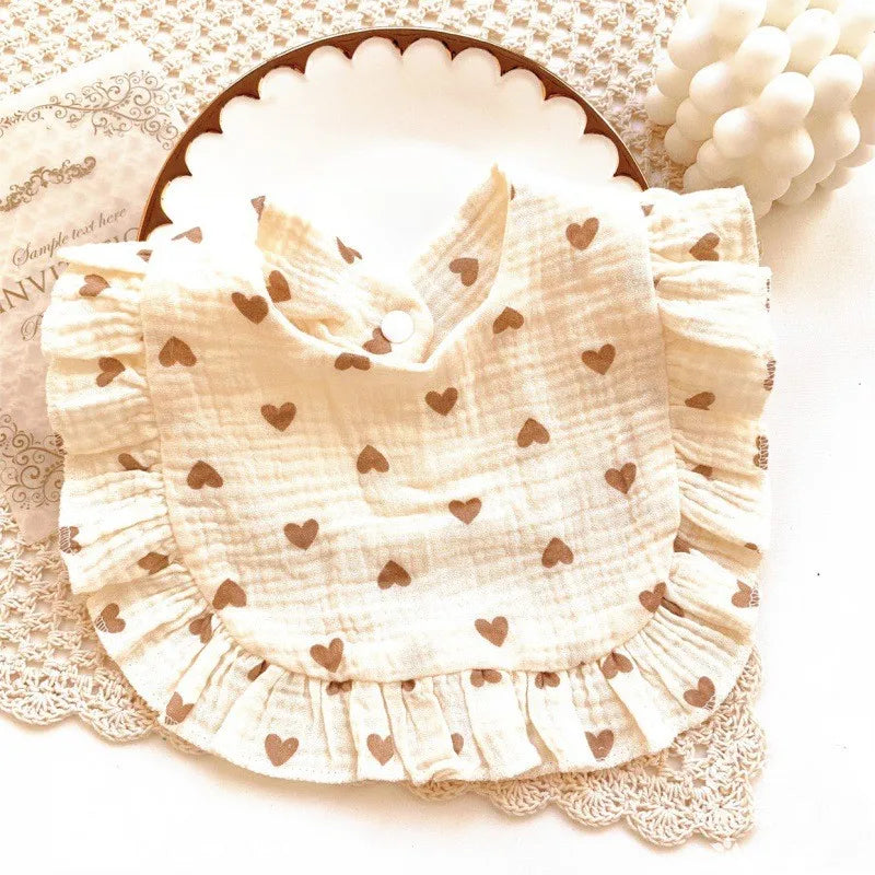 Cream-colored bib with brown heart patterns and ruffled edges displayed on a decorative plate. Background includes a lace doily and a candle shaped like rounded bubbles.