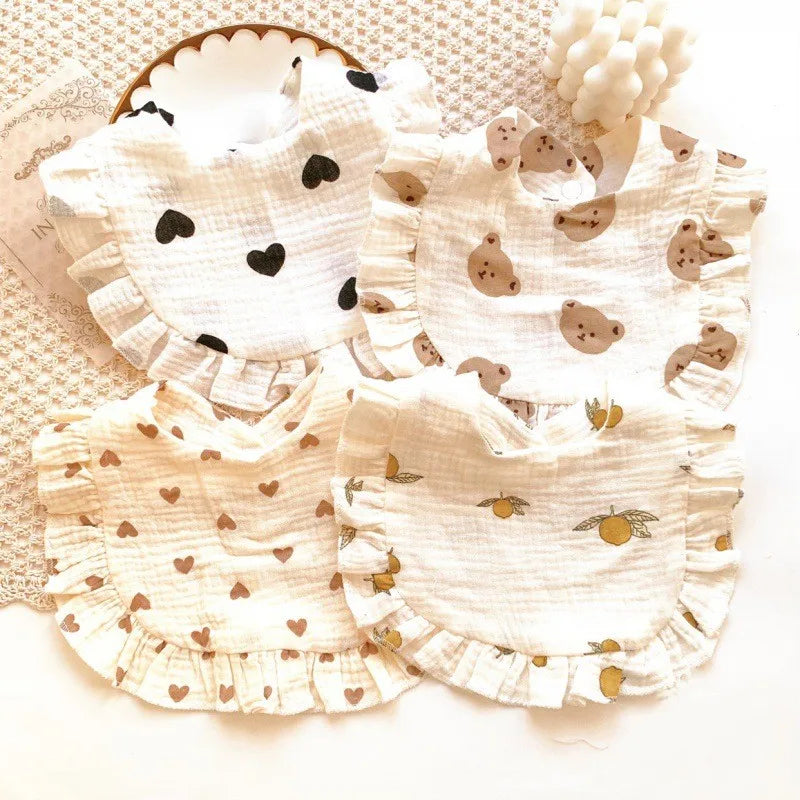 Four baby bibs with ruffled edges featuring different patterns: black hearts, bear faces, brown hearts, and oranges on a textured white background.