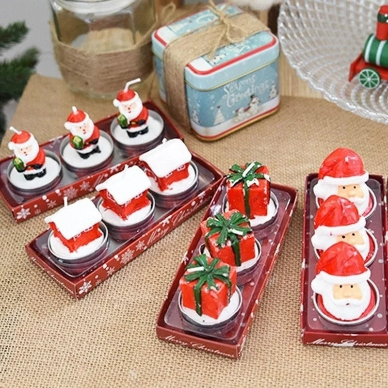 Decorative Christmas-themed candles in red boxes featuring Santa figures, houses with snowy roofs, and gifts with green ribbons, arranged on a burlap surface.