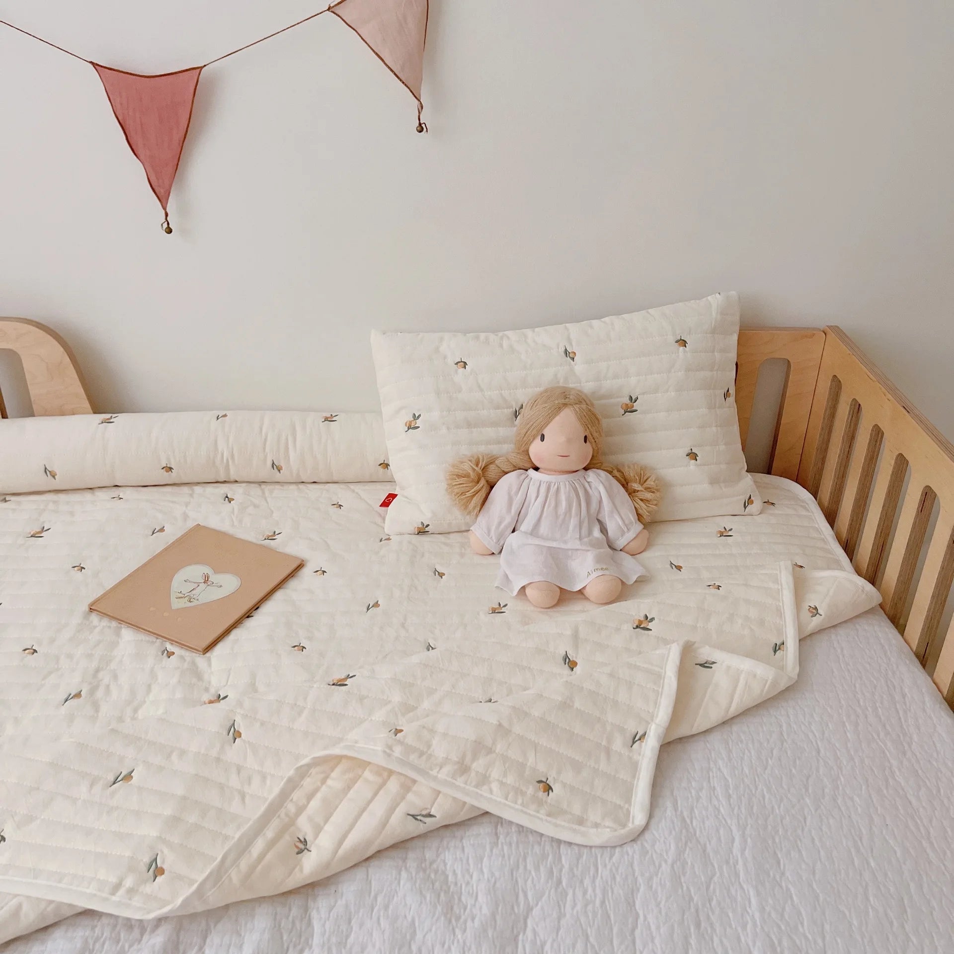 A cozy child's bed with a floral-patterned cream blanket and pillow, a doll in a white dress, a notebook, and a pastel bunting decoration on the wall.