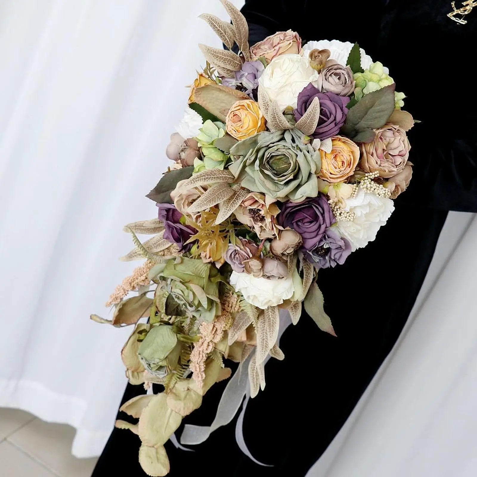 A cascading bouquet featuring roses in purple, peach, and cream, with greenery and textured leaves, held by a person in black attire against a white curtain backdrop.
