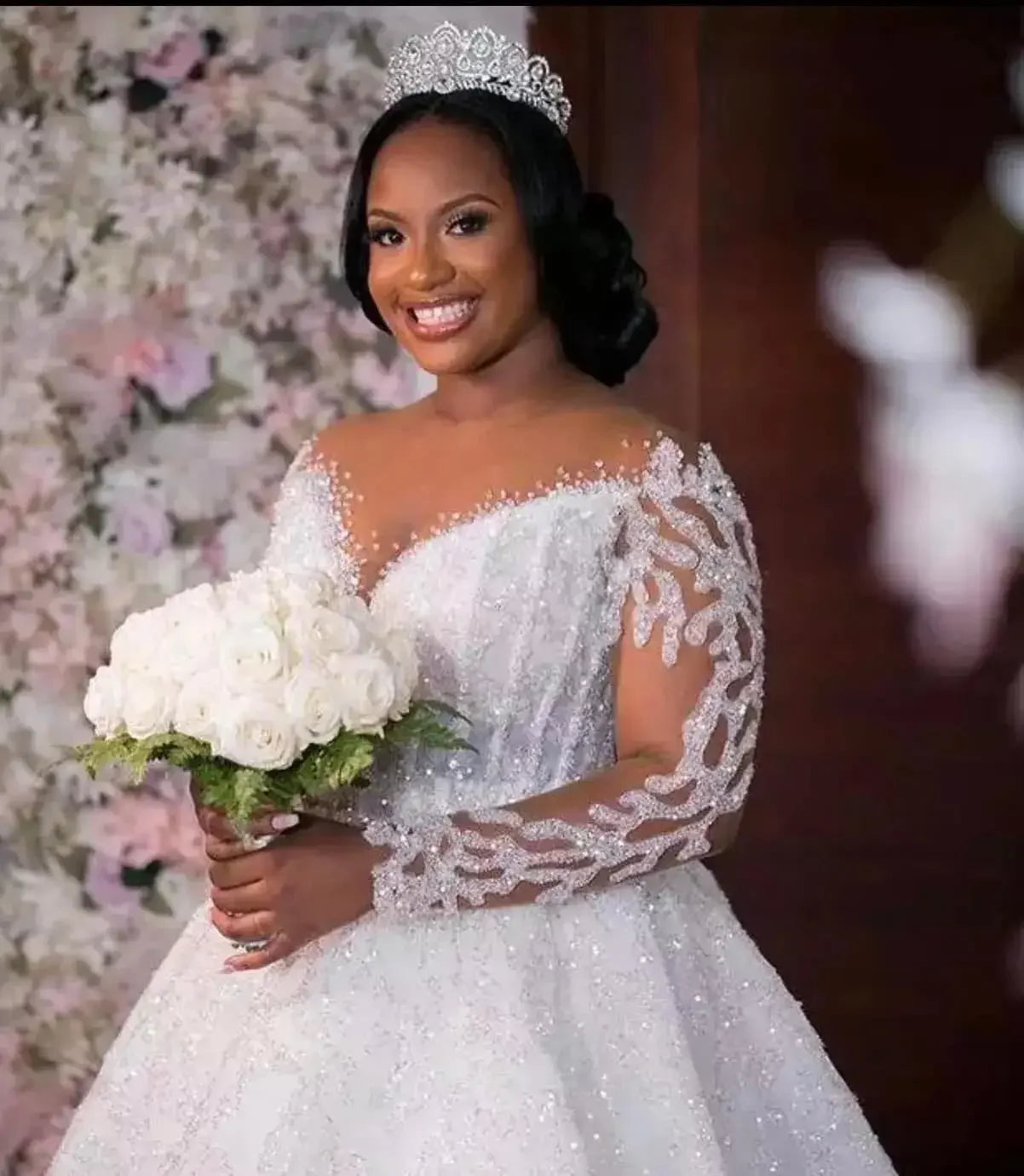 Bride in a sparkling white gown with lace details and a tiara, holding a bouquet of white roses. A floral background enhances the elegant setting.