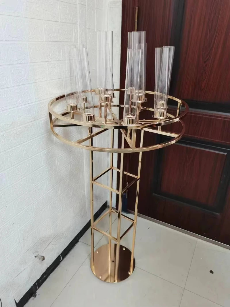 Tall, gold metal candle holder with a circular base and multiple clear glass tubes on top, standing on a tiled floor against a white textured wall and wooden door.