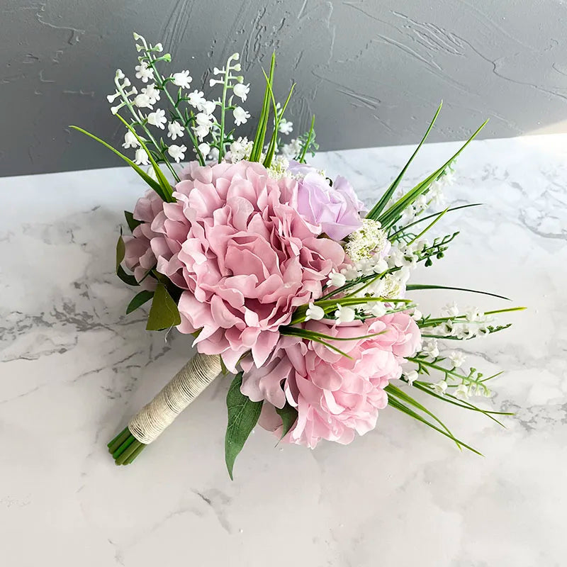 A bouquet of pink peonies, white lilies of the valley, and green foliage, wrapped with twine, lies on a marble surface against a gray textured wall.