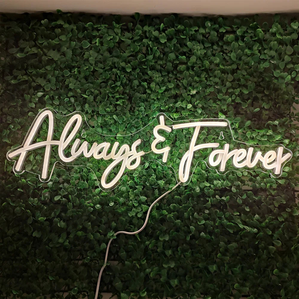 Neon sign reading "Always & Forever" glowing against a lush green foliage background. A white cord is visible below the sign.