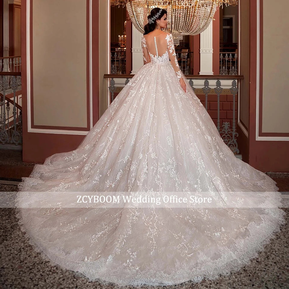 Bride in a lavish, lace-embroidered white wedding gown stands in an ornate room with a grand chandelier, looking back over her shoulder.