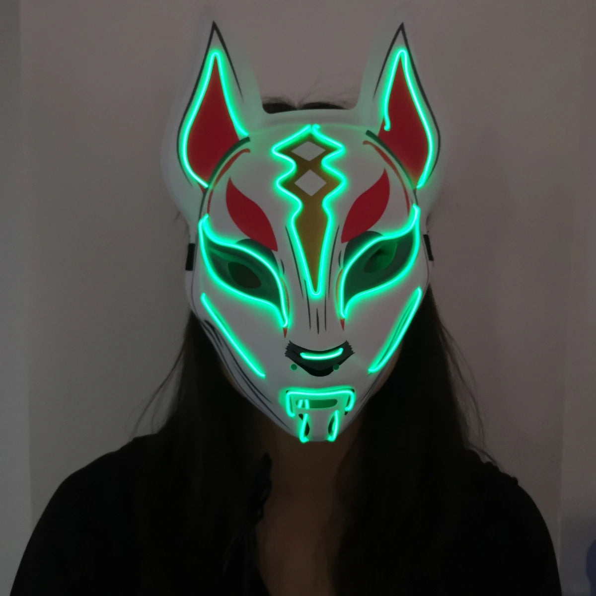 A person wearing a luminous fox mask with glowing green outlines and red accents, set against a dark background. The mask has pointed ears and intricate patterns.