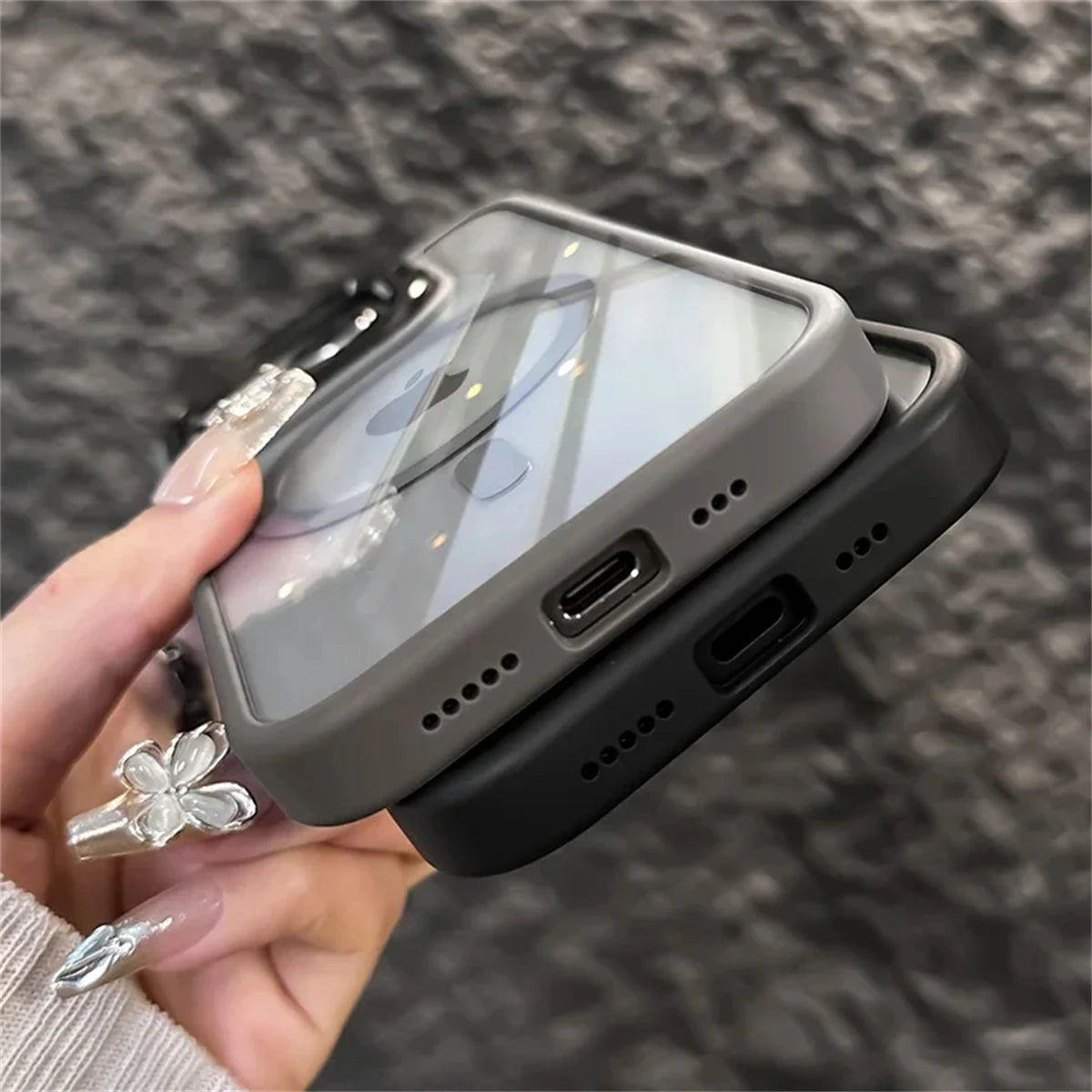 Two smartphones with gray and black cases, held by a person with manicured nails and a transparent clover ring, showing the charging ports at the bottom.