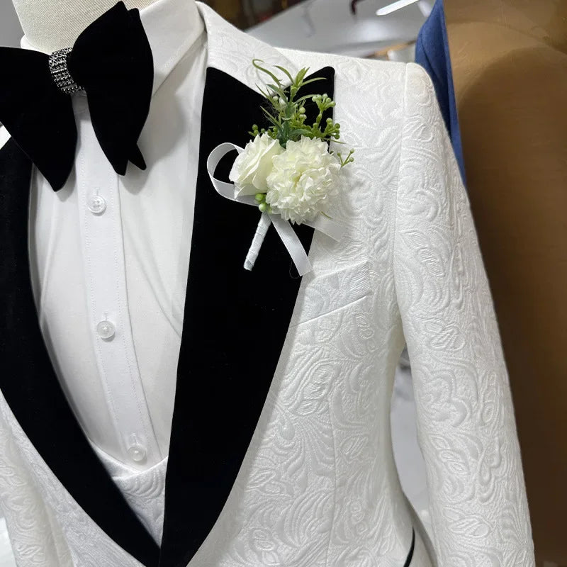 Close-up of a white embroidered tuxedo jacket with a black velvet lapel, worn with a white shirt and black bow tie. A white boutonniere with greenery is pinned on the lapel.