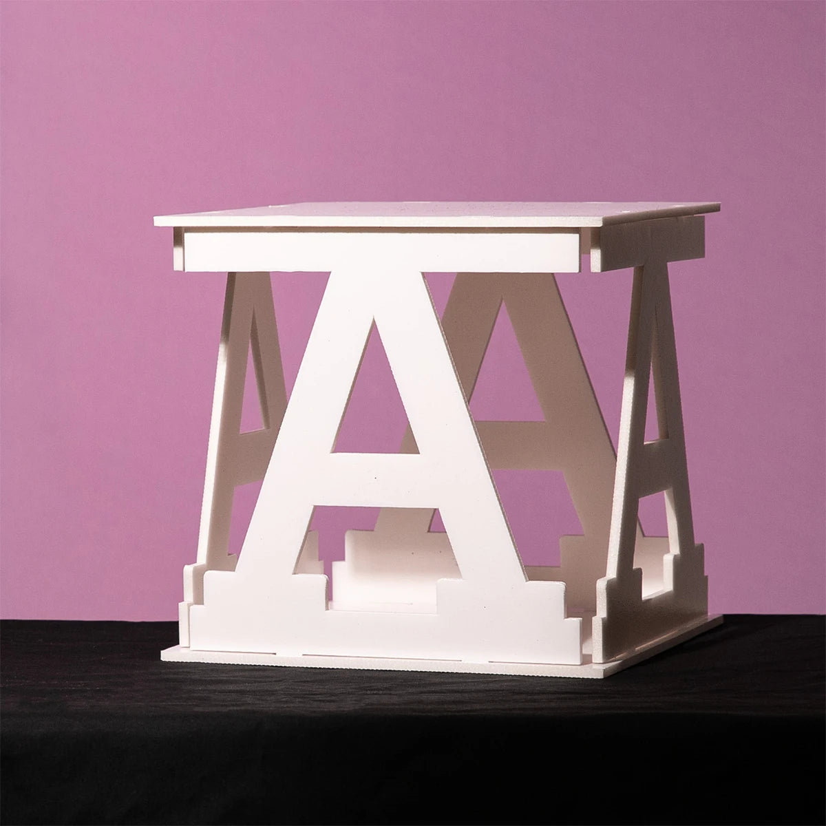 White sculptural object with an "A" shape design, placed on a black surface against a pink background.