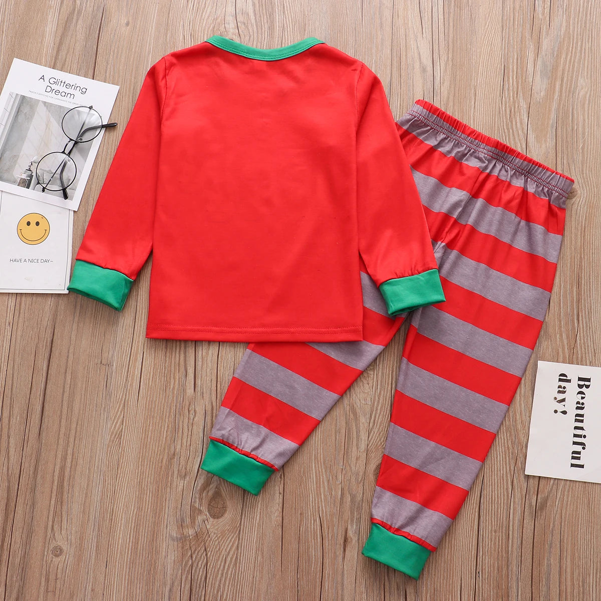 Red and green long-sleeve top with matching striped pants are laid out. The background has a wooden surface, with a pair of glasses and paper with text beside the outfit.