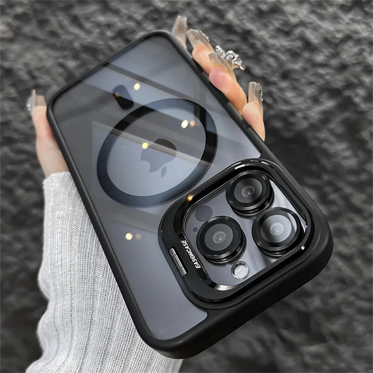 Close-up of a hand holding a smartphone with a black case, featuring a triple-lens camera. The reflection on the screen shows a logo. The hand has long, decorative nails.