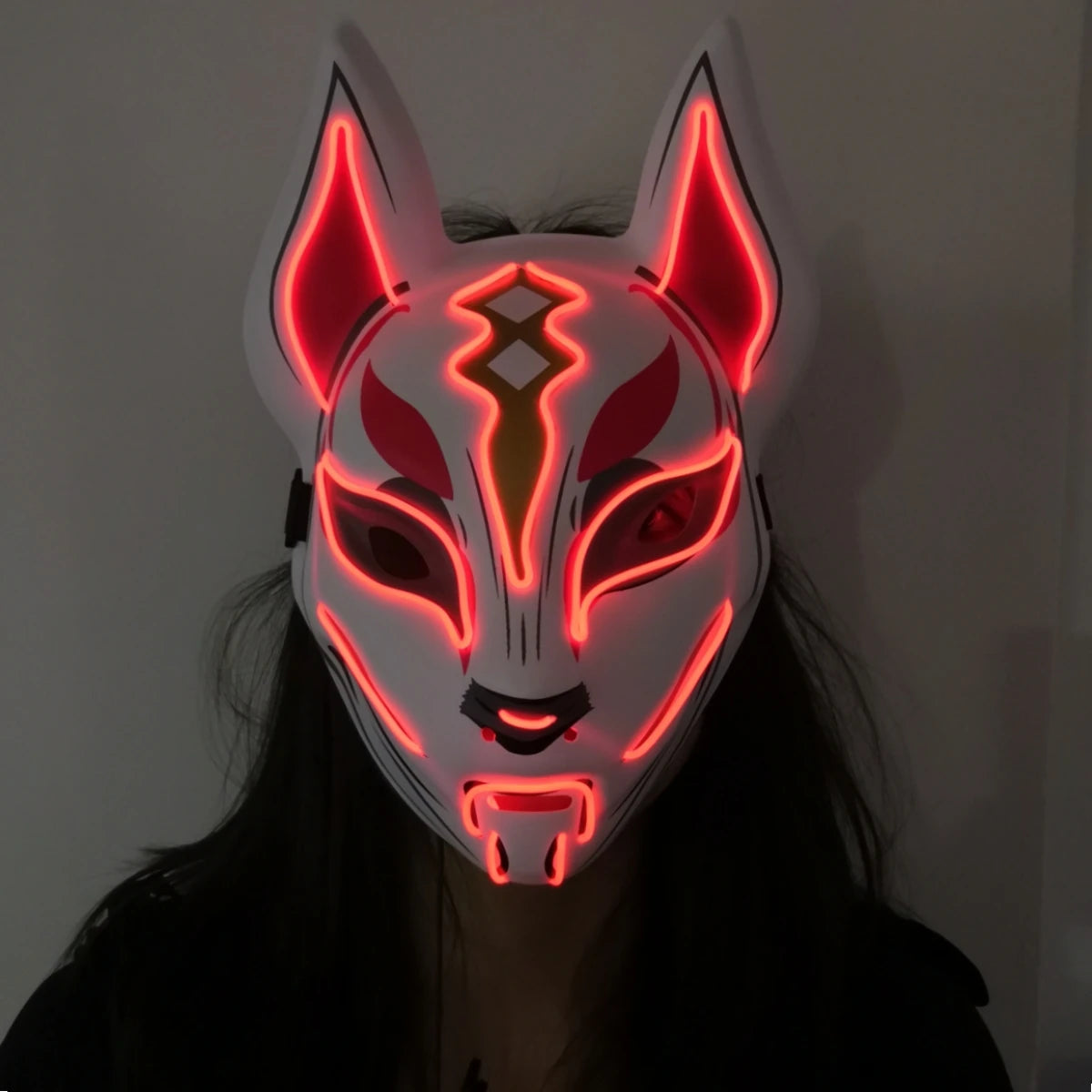 A person wears an LED fox mask glowing red, with intricate patterns and large ears. The mask covers the face completely, creating an otherworldly appearance against a dark background.
