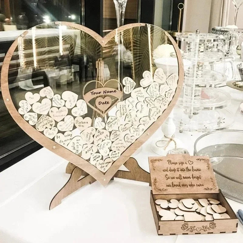 Heart-shaped guest book frame filled with signed heart tokens on a table beside an open box of blank hearts. Instructions encourage guests to sign and insert tokens.
