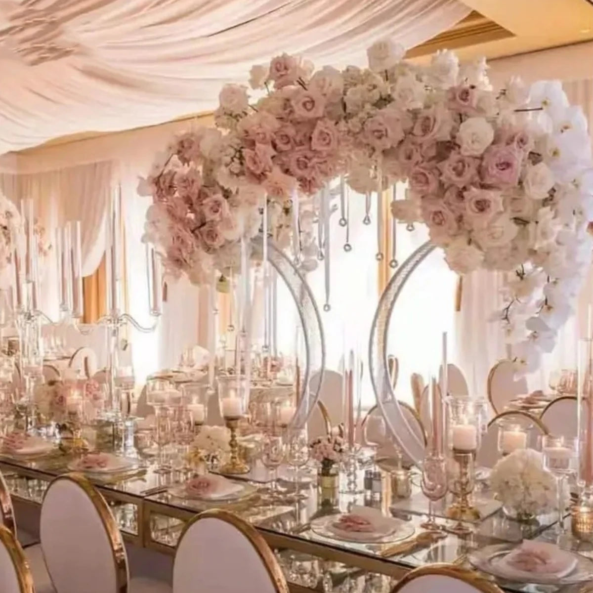 Elegant wedding table setting with mirrored surfaces, adorned with pink and white floral arrangements, tall candles, and gold-rimmed white chairs beneath draped fabric ceiling.