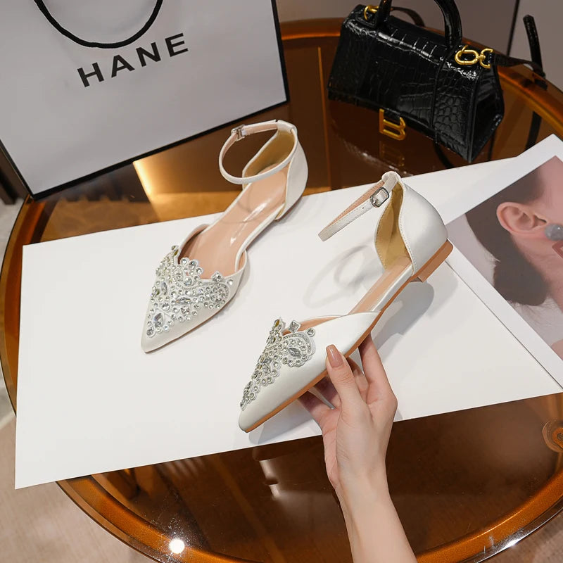 Elegant white pointed flats with ankle straps and rhinestone embellishments on a table. A hand holds one shoe, with a black handbag and paper bag in the background.