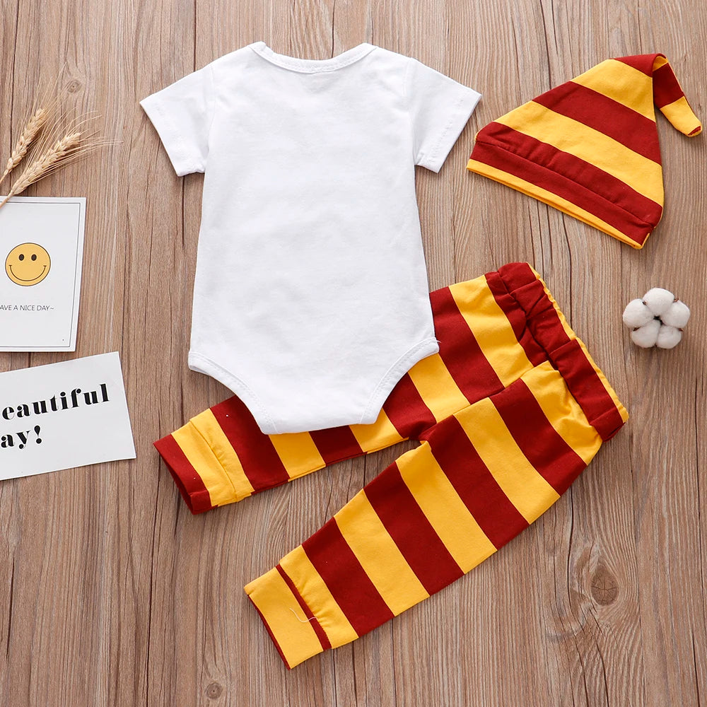 Baby outfit on wooden floor with white onesie, red and yellow striped pants, matching hat, and cotton ball. Smiley face and "Have a Nice Day" cards in the background.