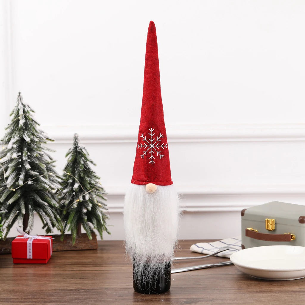 A holiday gnome wine bottle cover with a tall red hat featuring a white snowflake design, white beard, and round nose. Tabletop includes small snowy trees and a wrapped gift.