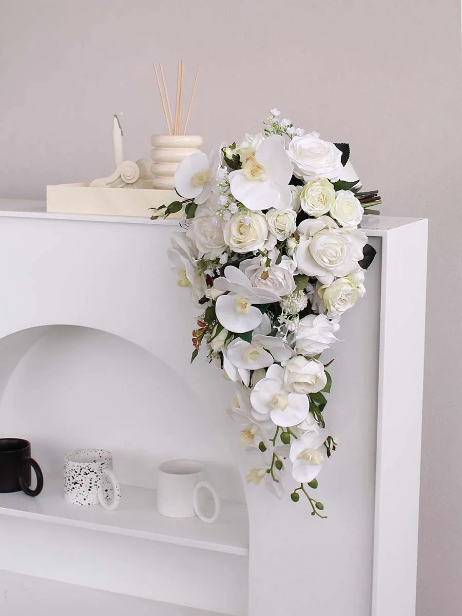A cascading arrangement of white flowers, including roses and orchids, decorates a white shelf with three mugs and an incense holder in a minimalist setting.