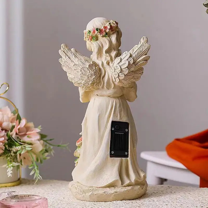 Back view of an angel figurine with wings and a floral crown, featuring a solar panel on its back. It stands on a table adorned with pink flowers and an orange cloth.