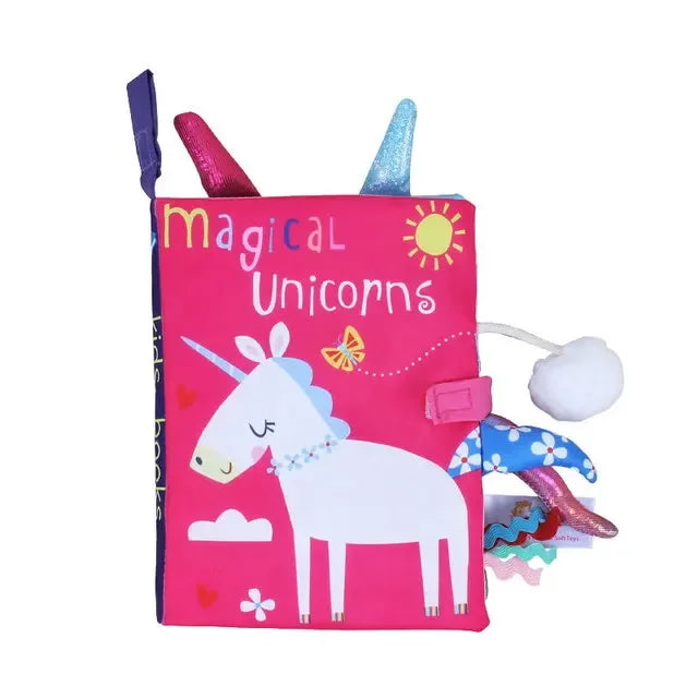 Bright pink cloth book titled "Magical Unicorns," featuring a white unicorn with colorful accents, a sun, clouds, and butterflies. Decorative tabs and textured elements are attached.
