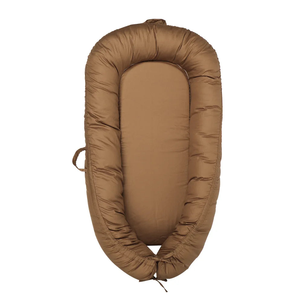 Brown baby lounger with padded edges and a soft, oval-shaped interior. It features a handle on one side and an adjustable tie at the bottom for size adjustment.