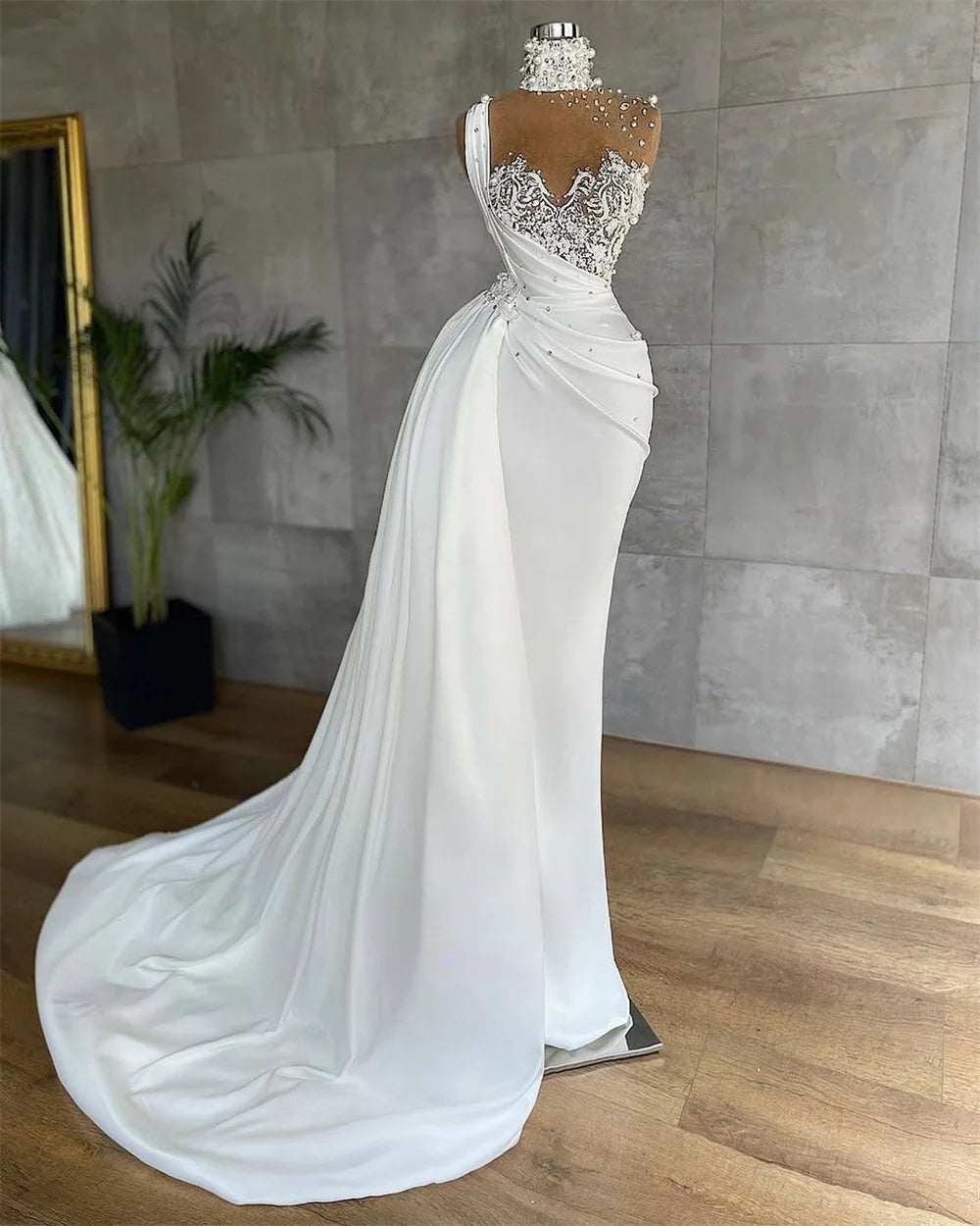 Elegant white gown on mannequin with intricate lace bodice and high neckline adorned with pearls. Draped fabric flows into a long train. Reflective mirror and plant in background.