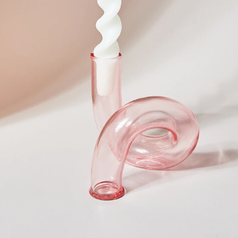 A pink curved glass candle holder with a sleek design holds a twisted white candle. The glossy surface reflects light, set against a neutral background.