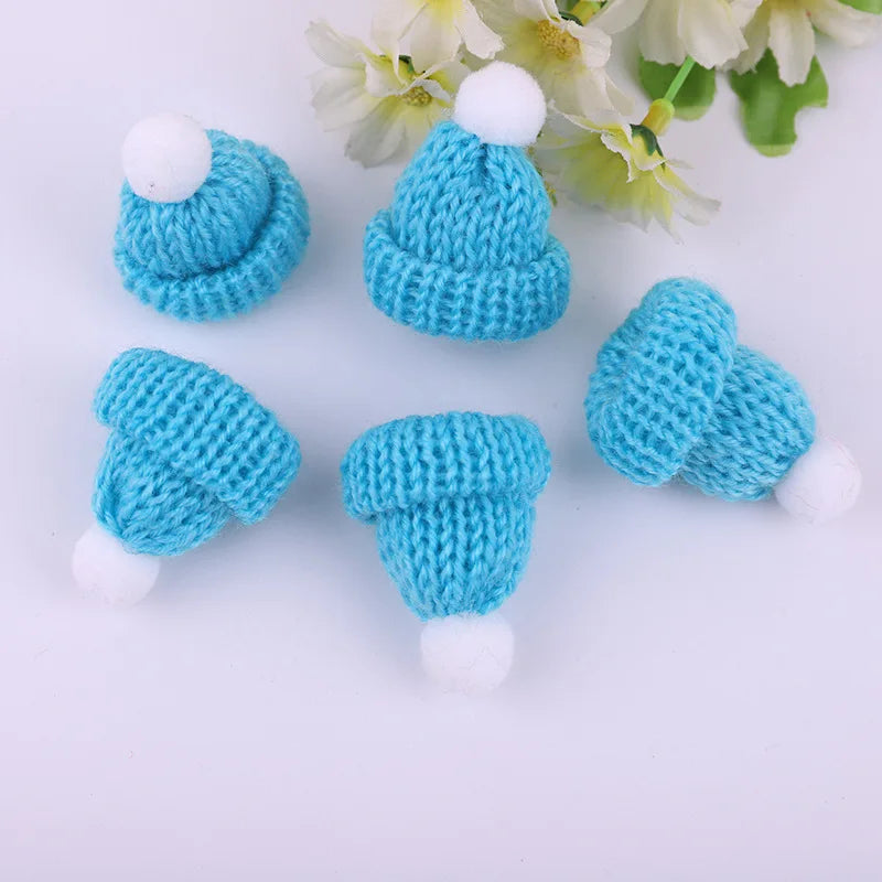 Five small blue knitted hats with white pom-poms are arranged on a white surface. Delicate white flowers with green leaves are in the background.