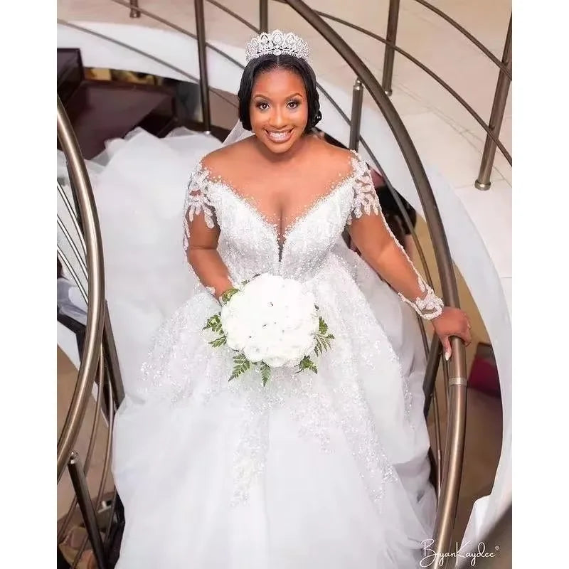 Bride in an ornate, sparkling white gown with lace sleeves stands on a curved staircase, holding a bouquet of white flowers. She wears a jeweled tiara and is smiling.
