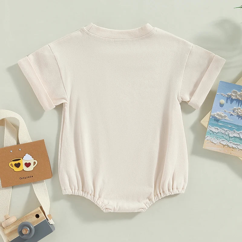Cream-colored baby onesie with short sleeves and elastic leg openings. Surrounding items include a toy camera with a strap, a card with teacup illustrations, and a beach painting.