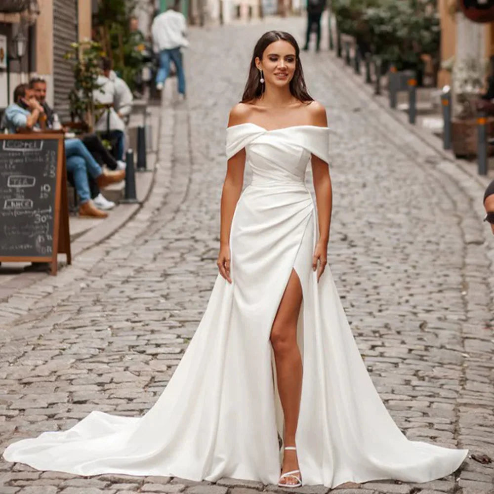A woman in an elegant off-shoulder white gown with a thigh-high slit walks down a cobblestone street lined with cafes and trees, exuding grace and style.