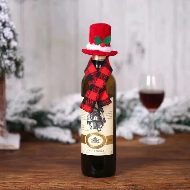 A wine bottle decorated with a red and black plaid scarf and a red top hat with holly. A glass of wine is in the background, with snowy decor around.