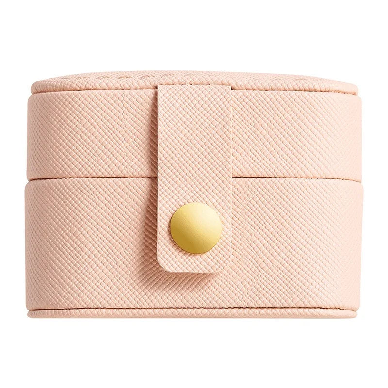 Compact beige cylindrical jewelry case with a textured surface and a gold button clasp. The lid is secured by a strap crossing the center.