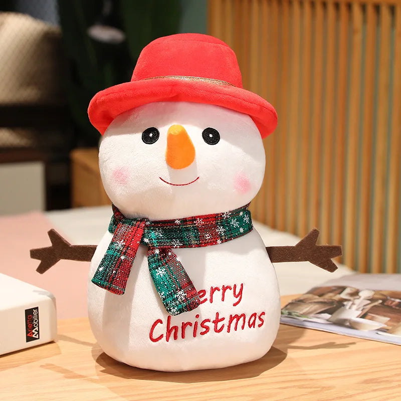 Plush snowman with red hat, orange nose, and embroidered "Merry Christmas" message. It wears a green and red scarf and has stick-like arms, sitting on a wooden surface.