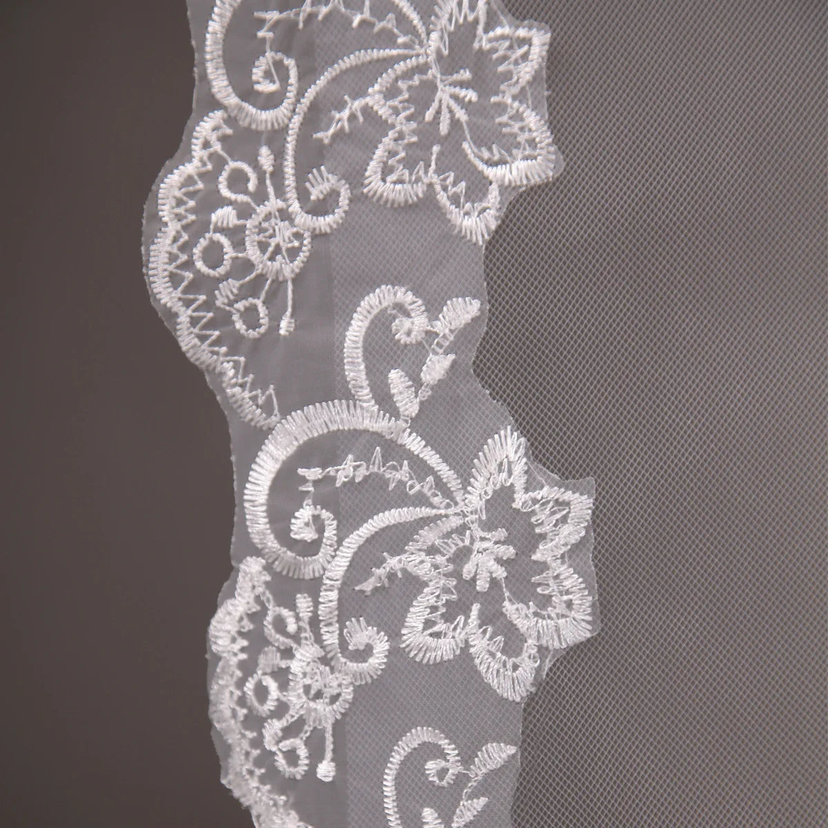 Close-up of intricate white floral lace embroidery on sheer fabric, featuring swirling patterns and delicate leaves, set against a plain background.