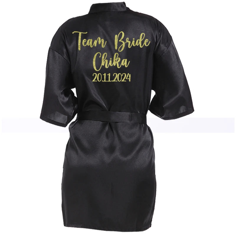 Black satin robe with gold script "Team Bride Chika 2011.2024" on the back. The robe features short sleeves and a matching belt around the waist.