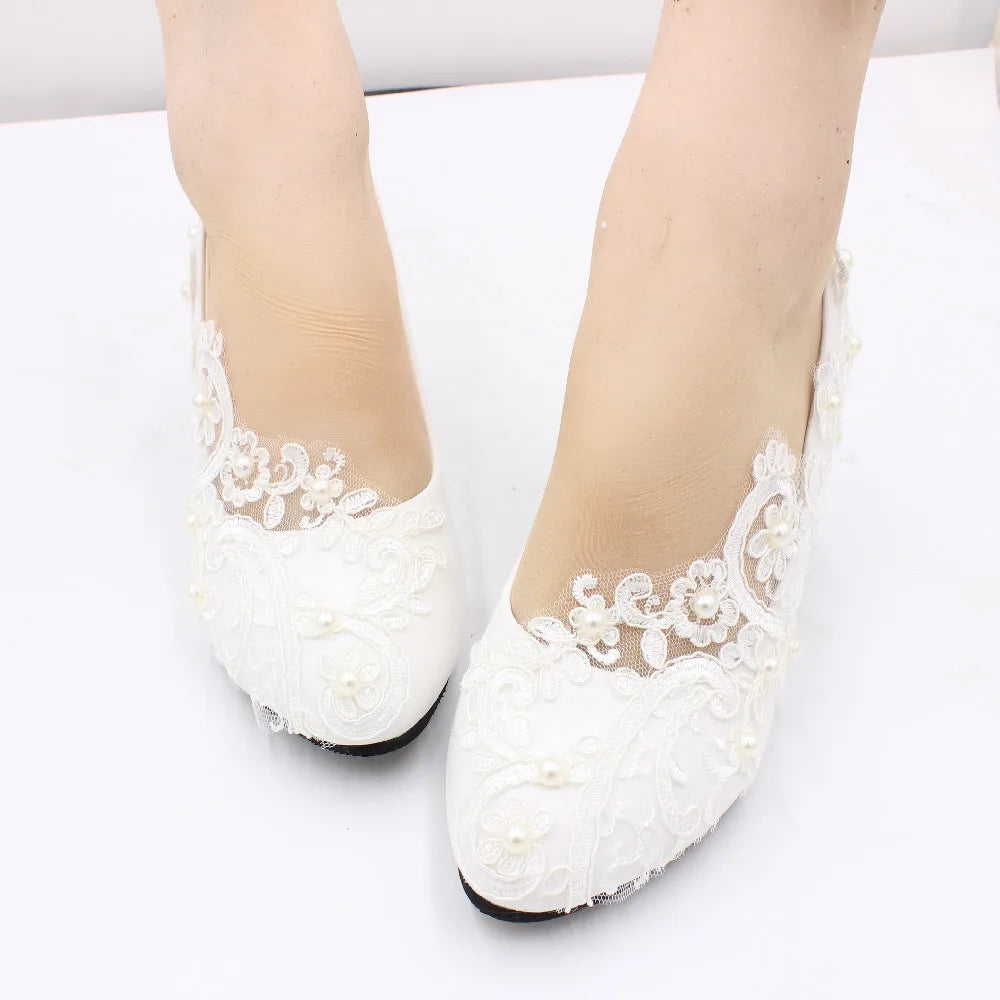 Close-up of white lace bridal shoes with intricate floral patterns and pearl accents. The shoes are worn on light-skinned feet, resting on a plain white surface.