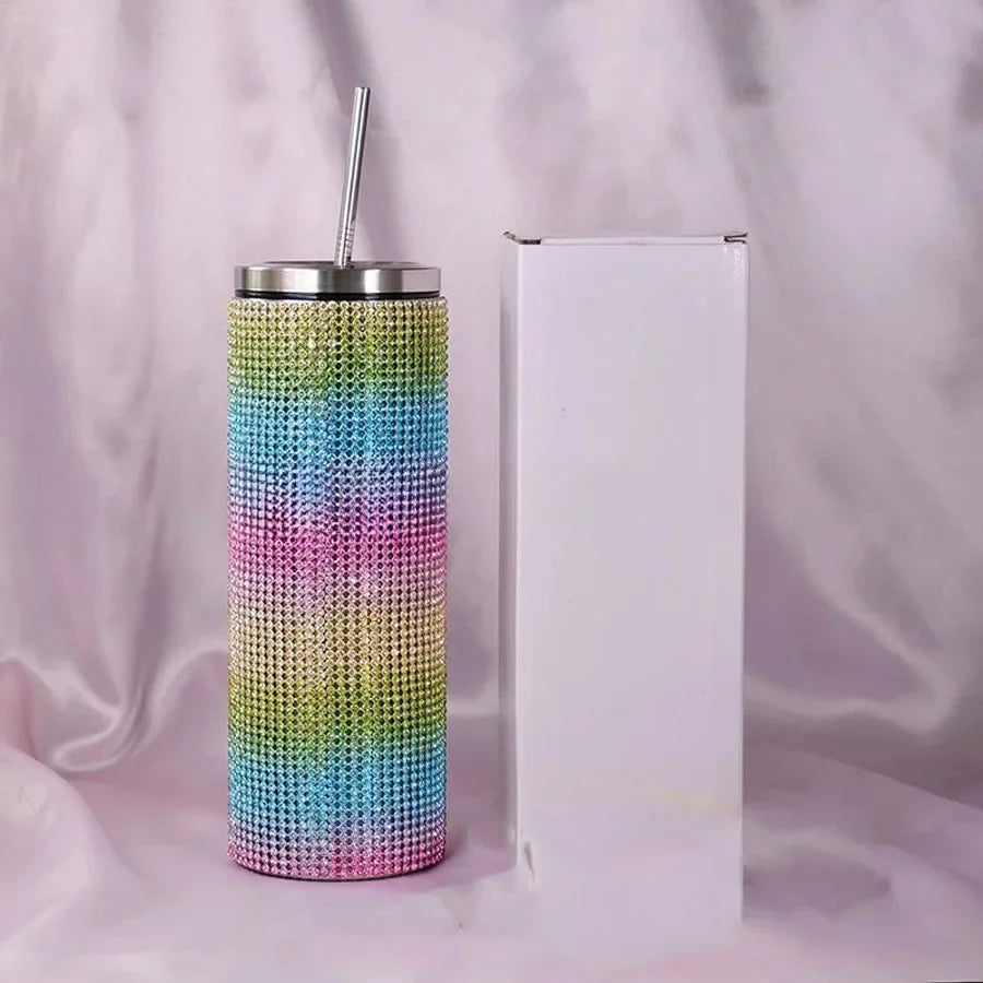 A multicolored, rhinestone-covered tumbler with a stainless steel lid and straw sits beside a plain white box against a soft, blurred background.