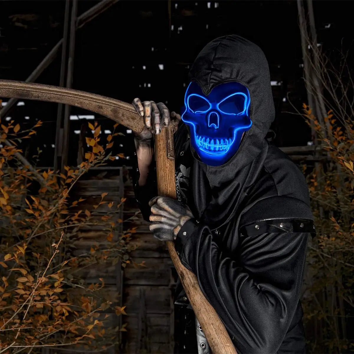 A person in dark clothing holds a wooden staff, wearing a glowing blue skull mask. The background features a dimly lit, overgrown wooden structure with autumn leaves.
