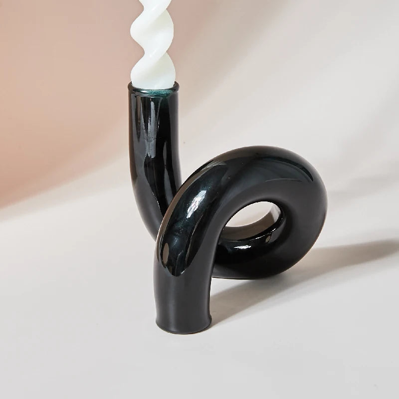 A black ceramic candle holder with a modern, curvy design holds a white twisted candle. The holder's sleek finish contrasts against the soft, neutral background.