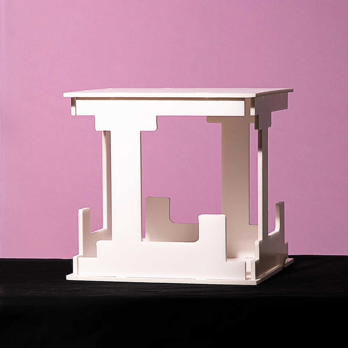 Minimalist white cubic table with cut-out geometric shapes, set against a pink background. The table is on a dark surface, highlighting its clean lines and modern design.