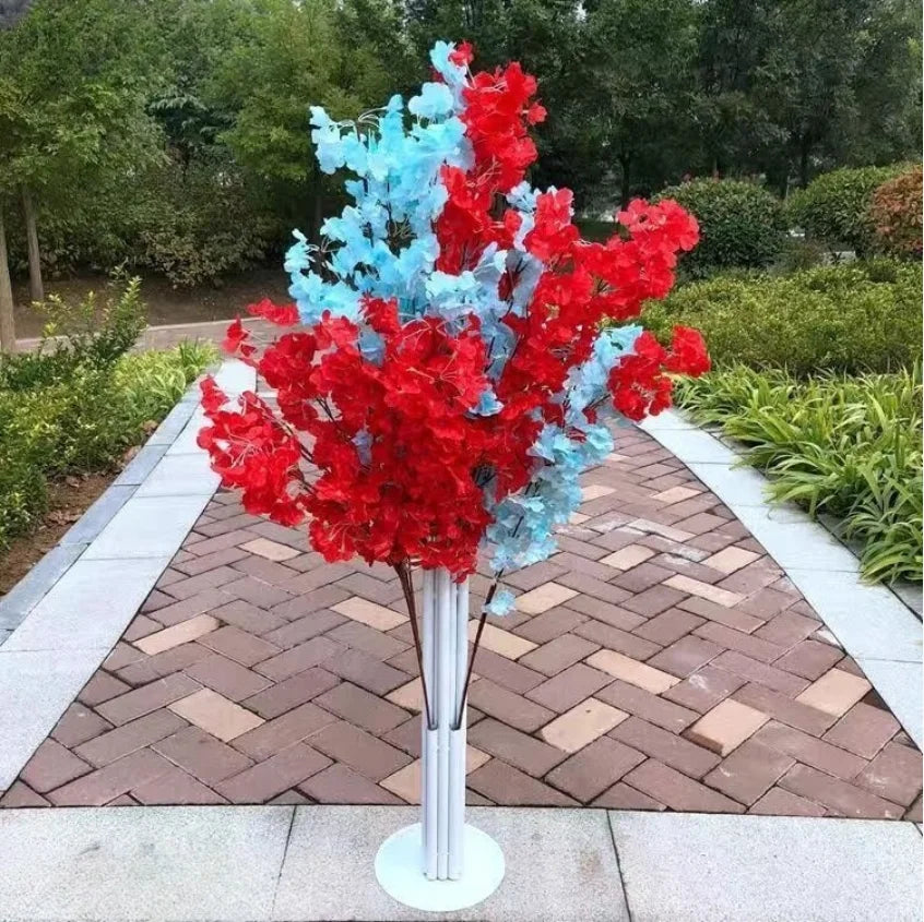 A tall vase with vivid red and soft blue artificial flowers stands on a brick pathway, surrounded by greenery in a garden setting.