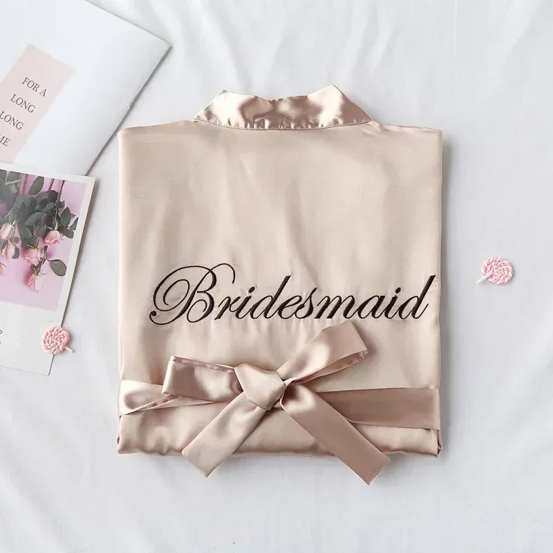 Beige satin robe folded neatly with "Bridesmaid" embroidered on the back. A matching bow is tied at the waist. Pink candies and a floral card are nearby on a white background.