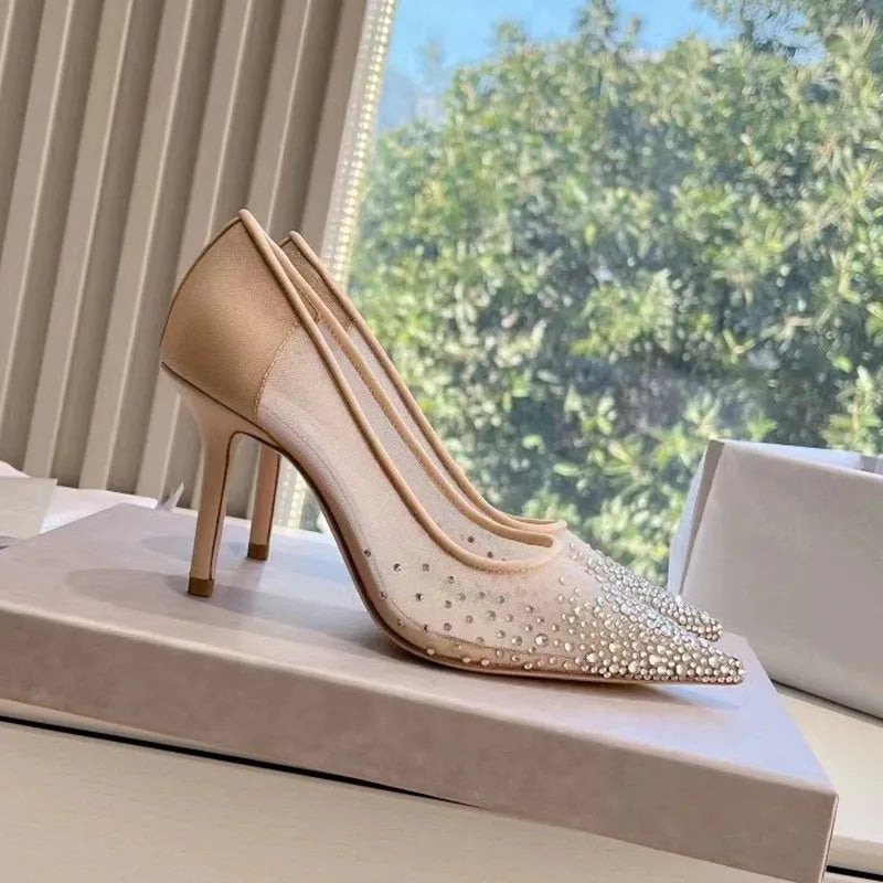 Elegant stiletto heel with a pointed toe, adorned with sparkling rhinestones, sits on a box. Background shows a window with a view of green foliage outside.