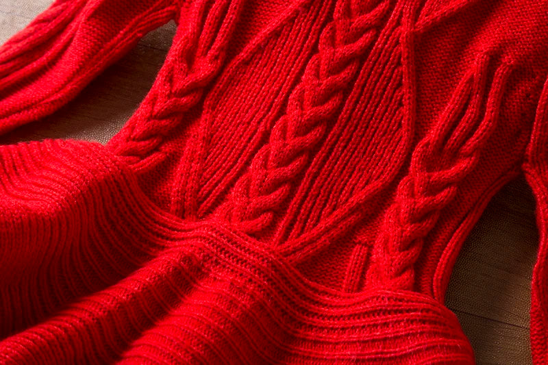 Close-up of a red knitted sweater with intricate cable patterns on the front. The fabric texture is detailed and visible, showcasing the craftsmanship and vibrant color.