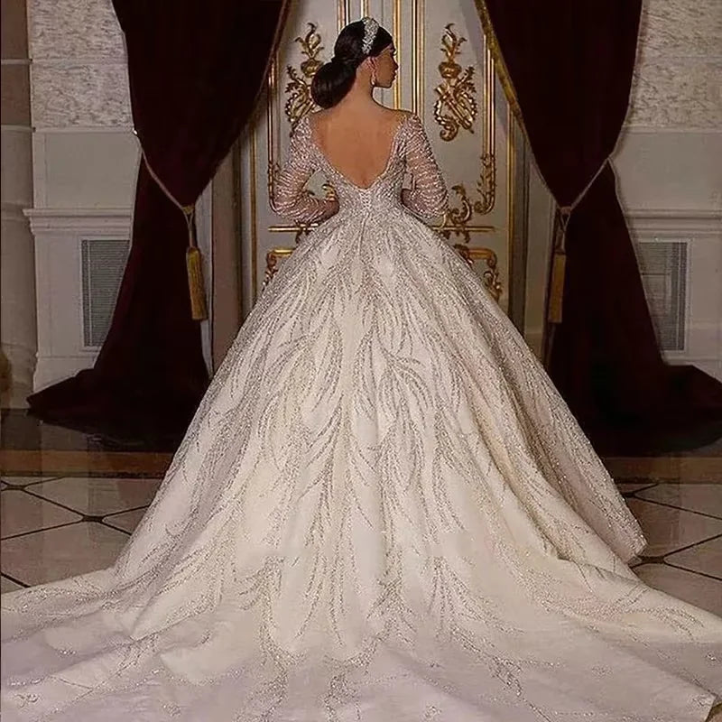 Elegant bride in a lavish white gown with intricate lace, standing before ornate golden doors. The dress features a long train and deep V-back, with dark curtains framing the scene.