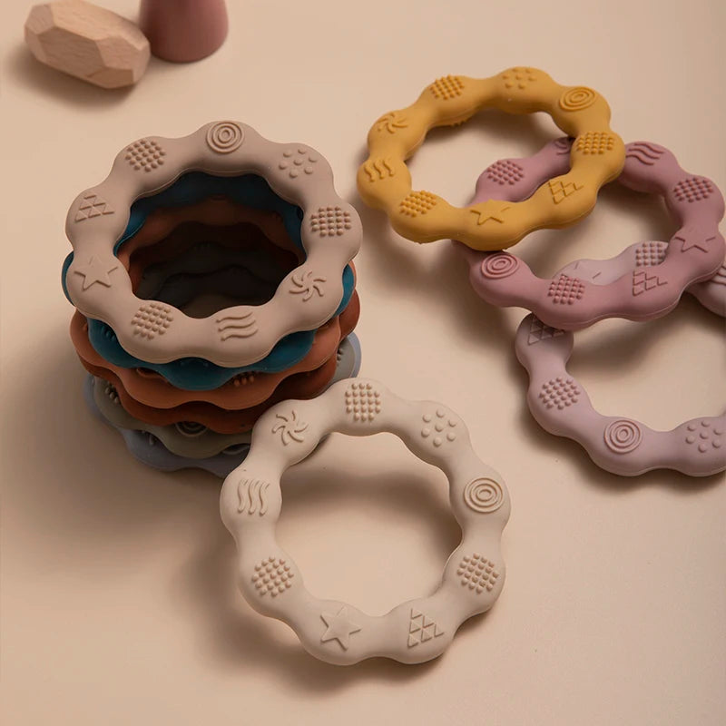 Stack of textured, round silicone teething toys in pastel colors, including beige, yellow, and pink, arranged on a light surface. Each ring has various embossed patterns.