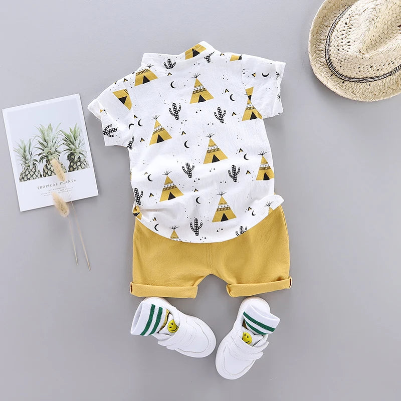 Toddler outfit with a white teepee and cactus print shirt, yellow shorts, white sneakers with green-striped socks, a straw hat, and a pineapple art print on a gray background.