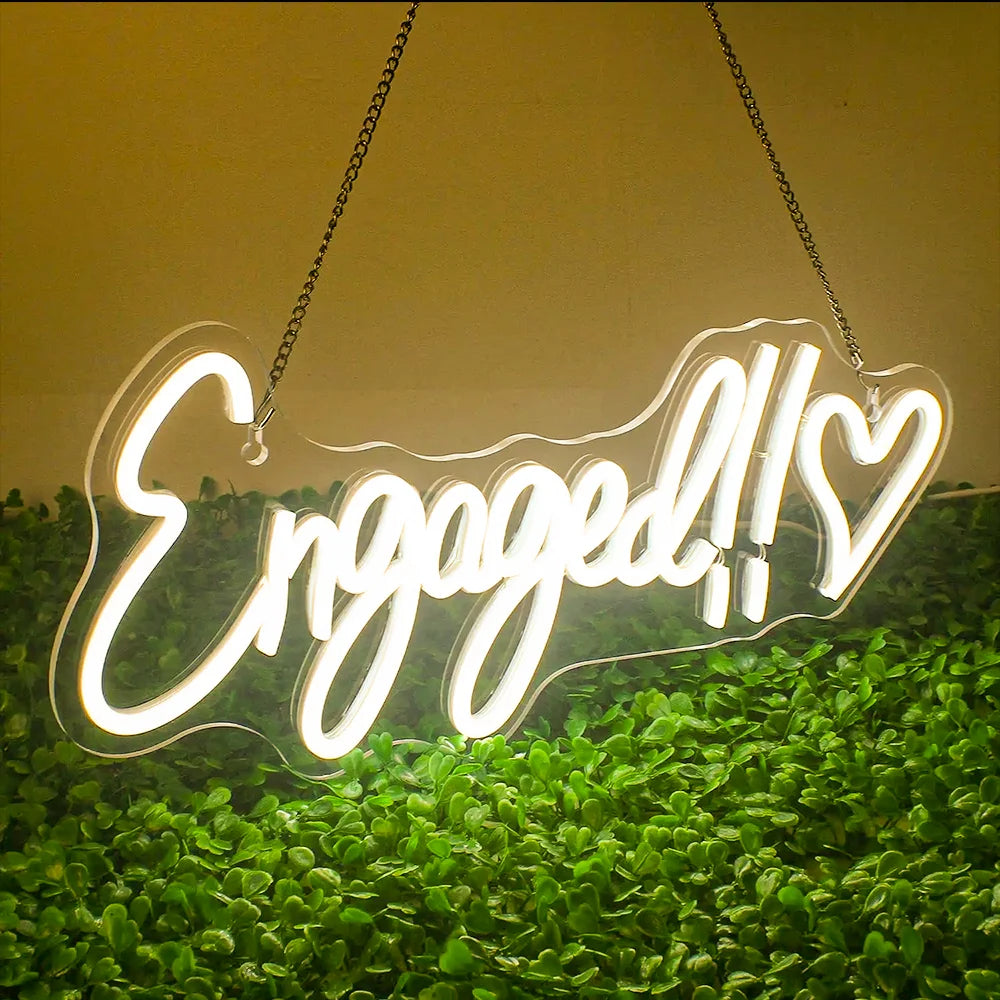 A neon sign reading "Engaged!!" with a heart symbol, hanging on chains. The sign glows brightly against a backdrop of lush green leaves.
